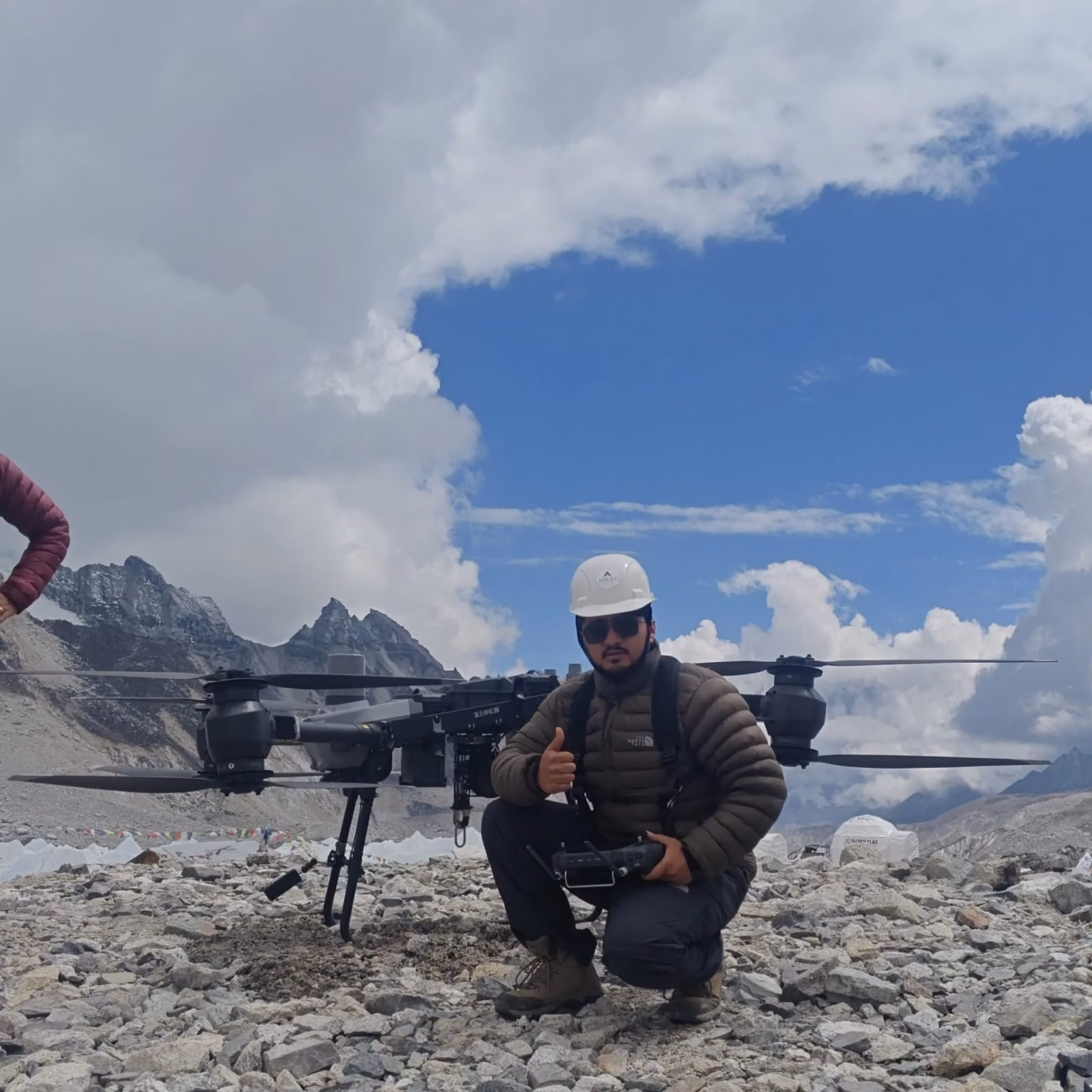 An operator crouched beside a survey drone on a glacial moraine, snow peaks behind