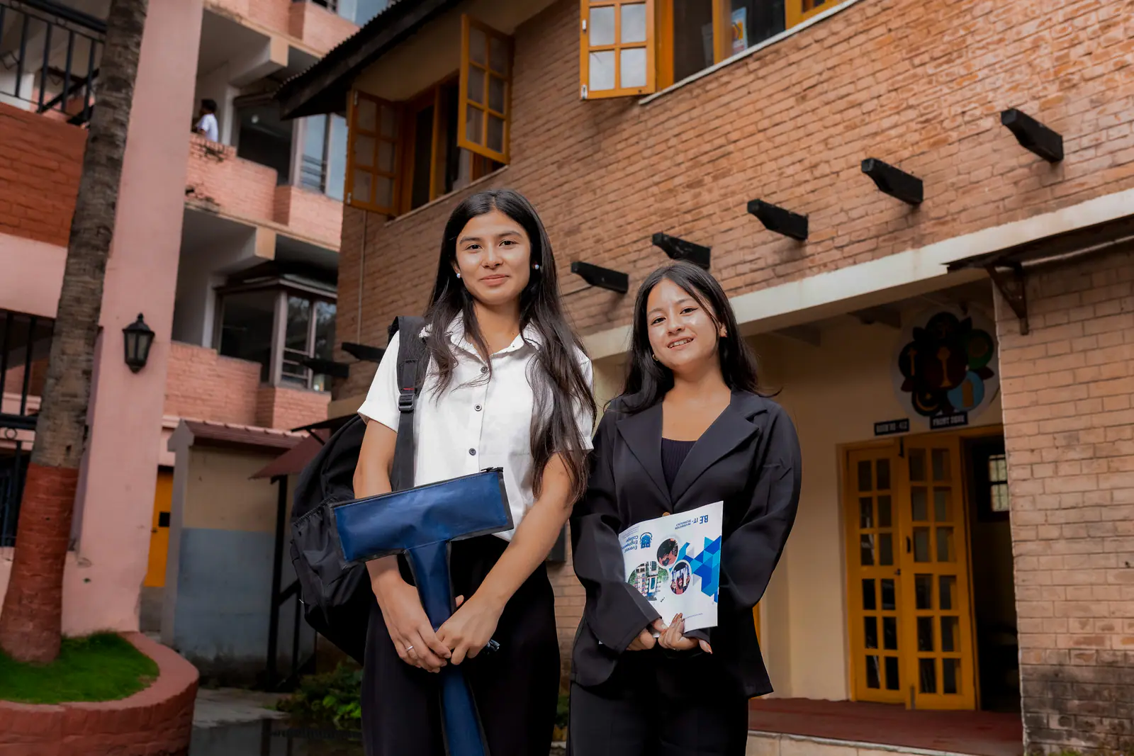 Two students in the paved courtyard of the red-brick teaching block, its yellow doors and awnings behind