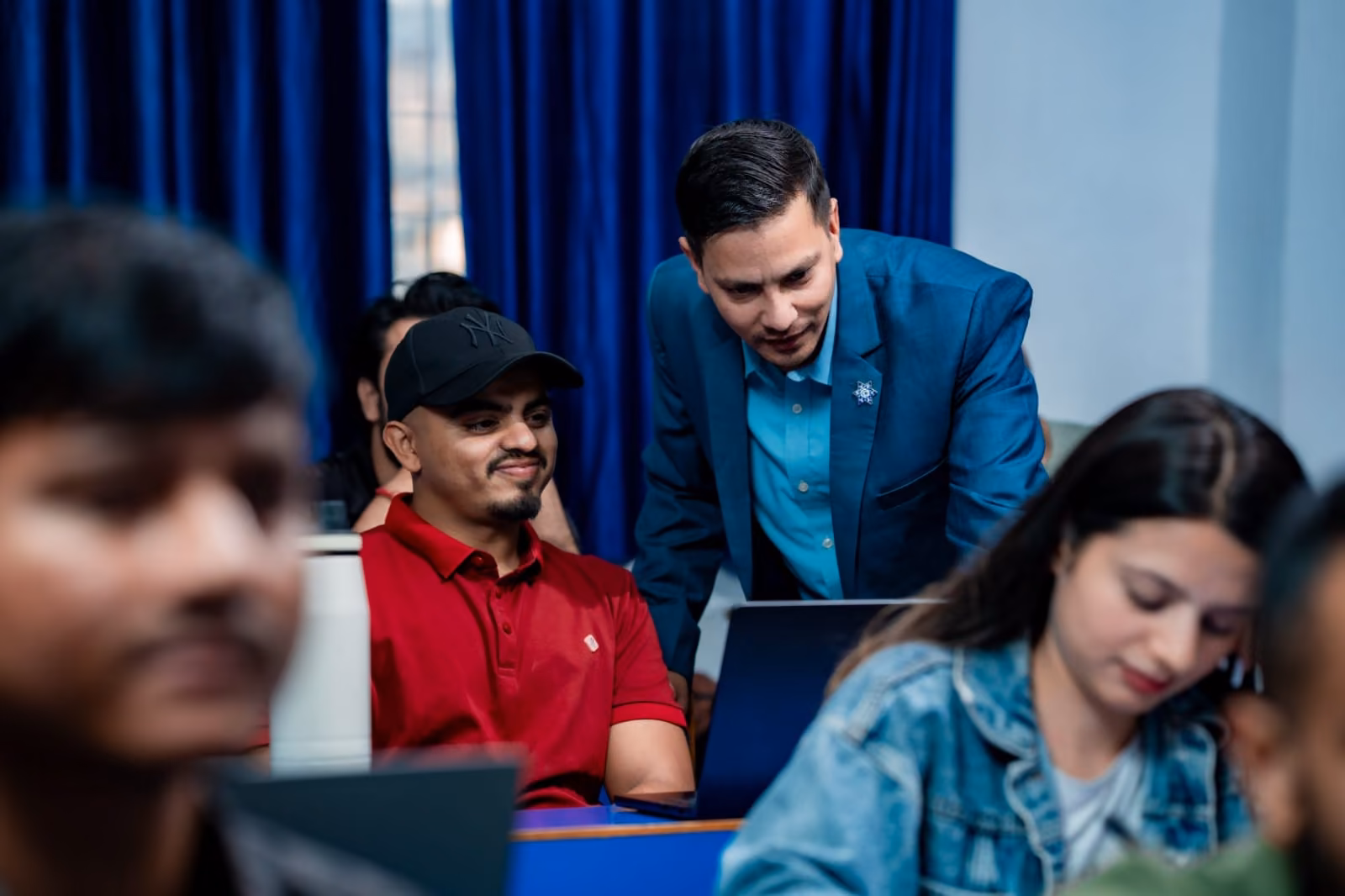 A tutor going round students at their laptops in a class, blue curtains drawn behind