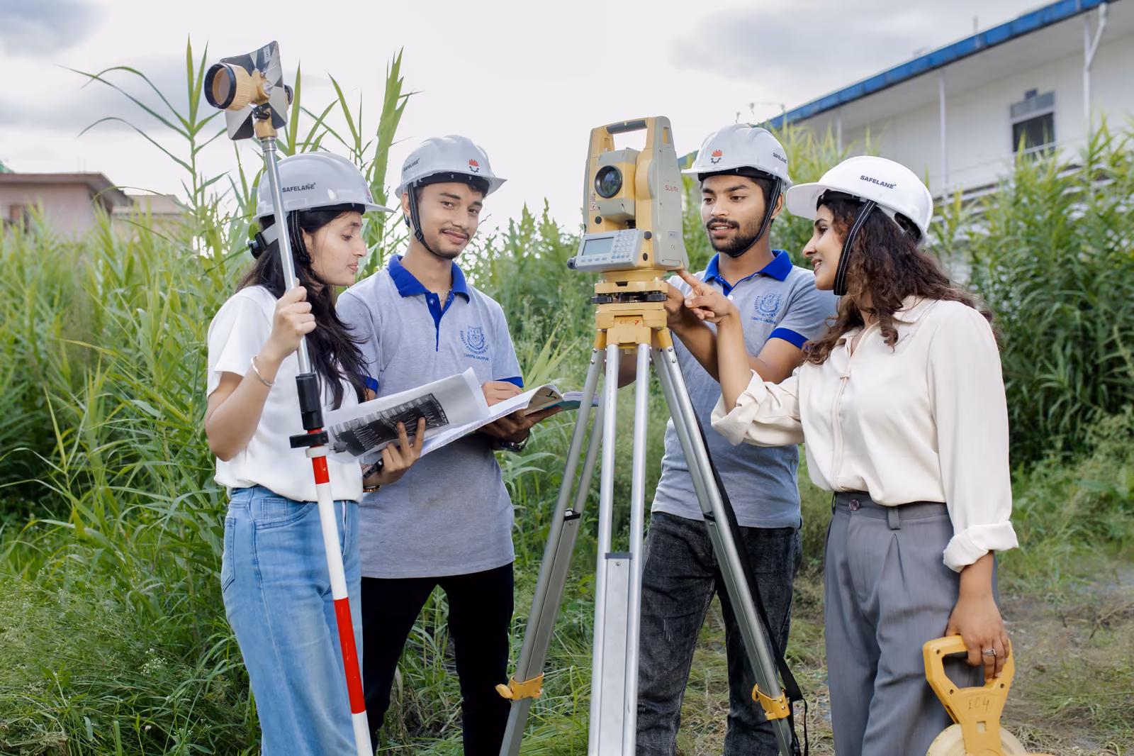 Four students in hard hats setting up a total station in long grass, one holding the levelling staff