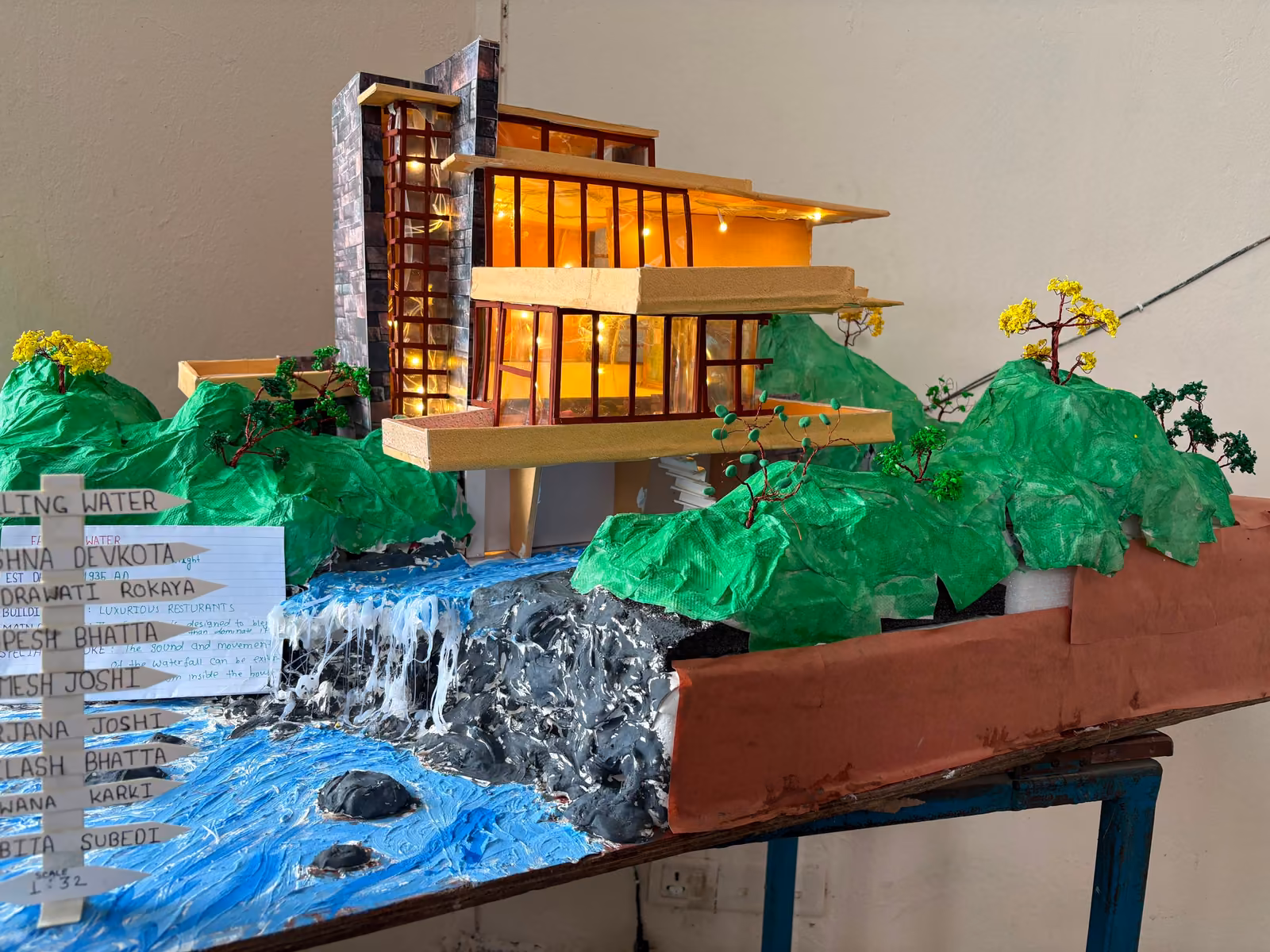 A student model of a timber and glass house on a green hillside, with a painted waterfall running past it