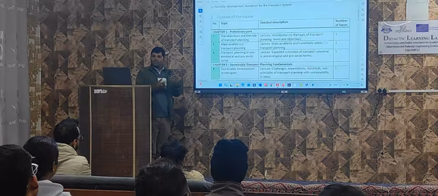 A speaker beside a wall-mounted screen in a panelled lecture hall, an audience seated in front of him