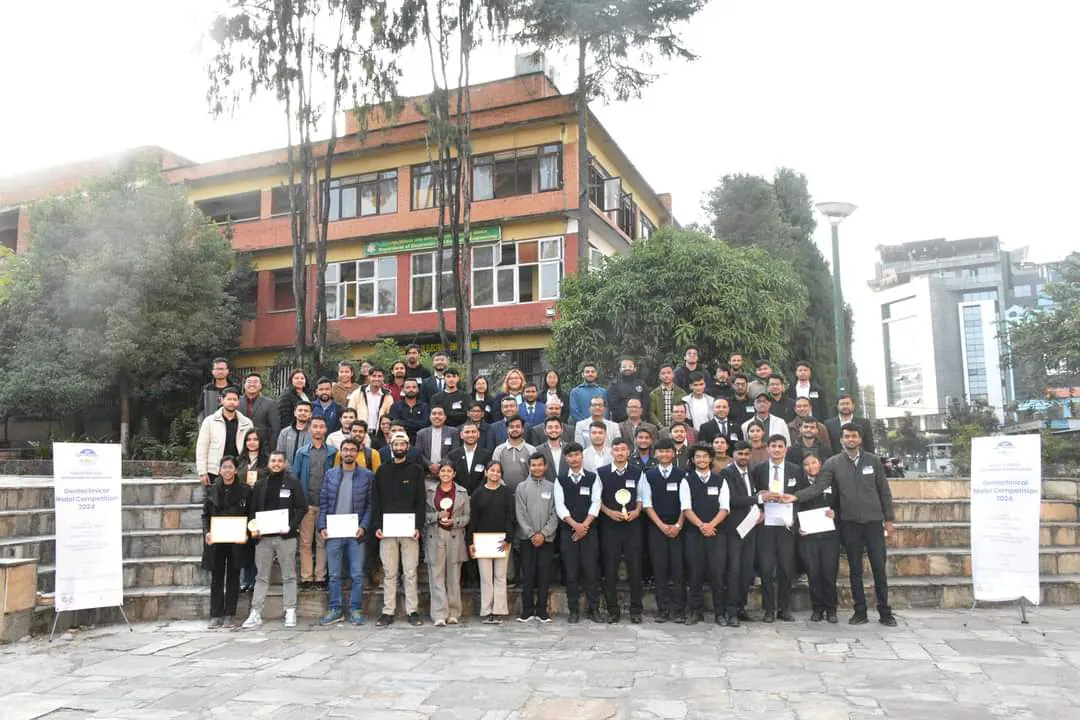 Around eighty students and staff standing in rows outside a long red-and-cream teaching block, trees behind