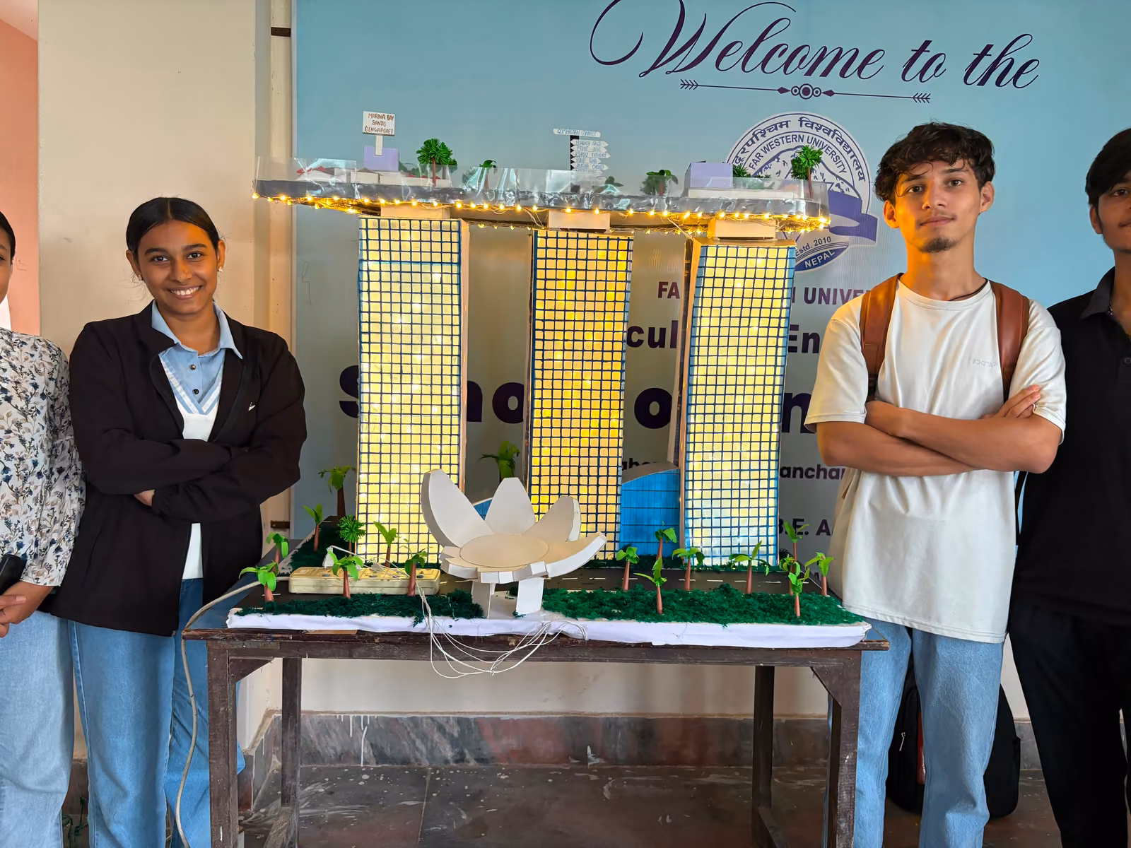 Two students either side of a table holding a tall yellow tower model with green landscaping around its base