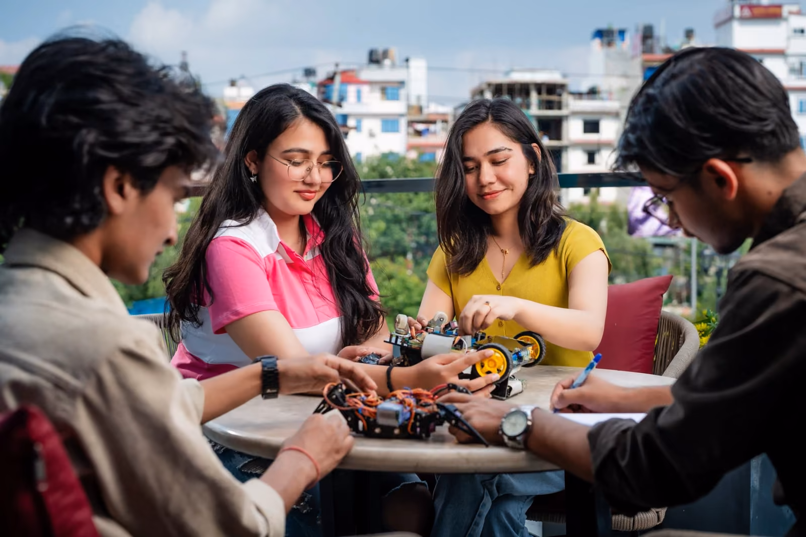 Four students round a table assembling a wheeled robot on a rooftop, the city behind