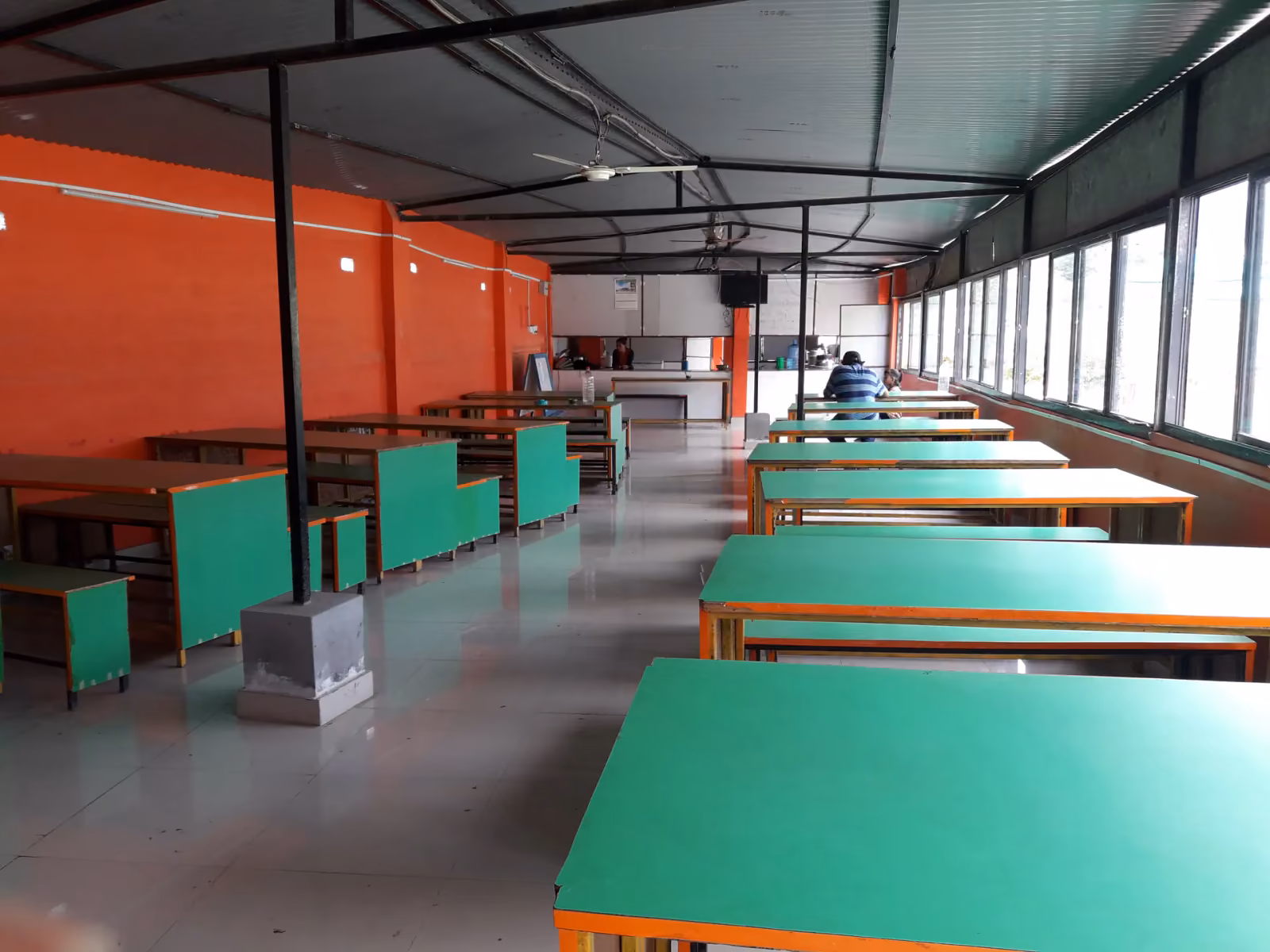 A long canteen of green-topped tables under a corrugated roof, windows down one side