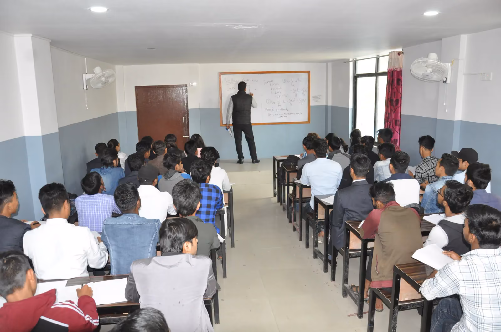 A full lecture room seen from the back, the lecturer writing at the whiteboard