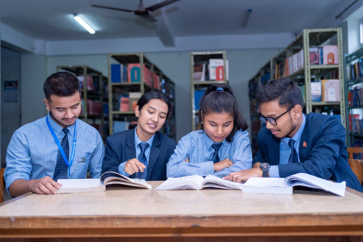 Four students reading open textbooks at a library table, the shelves behind them