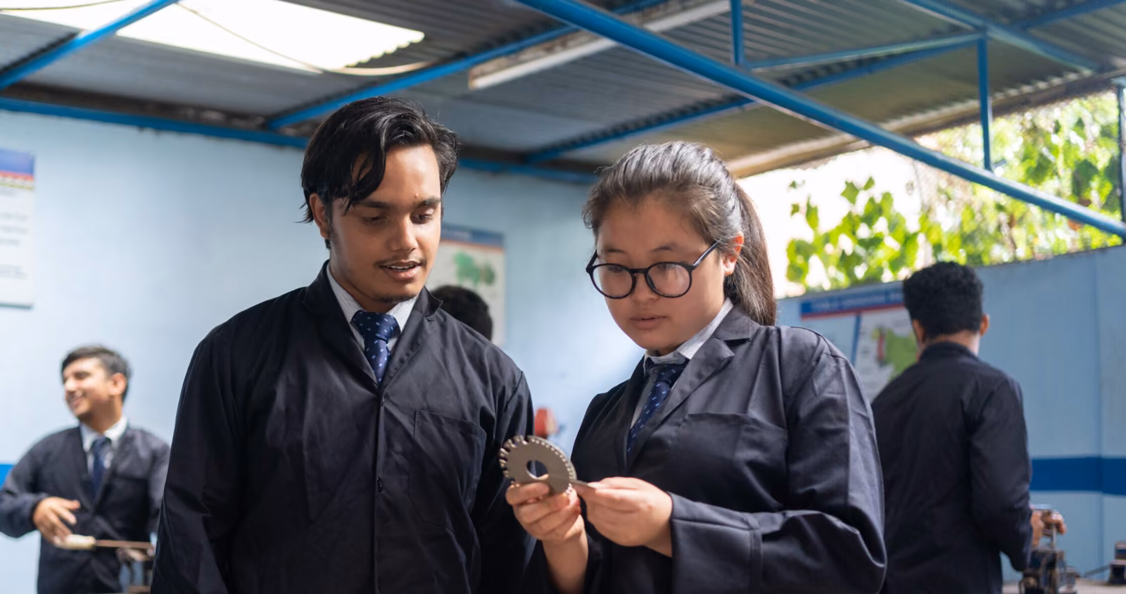 Two students in lab coats examining a toothed metal part in the workshop