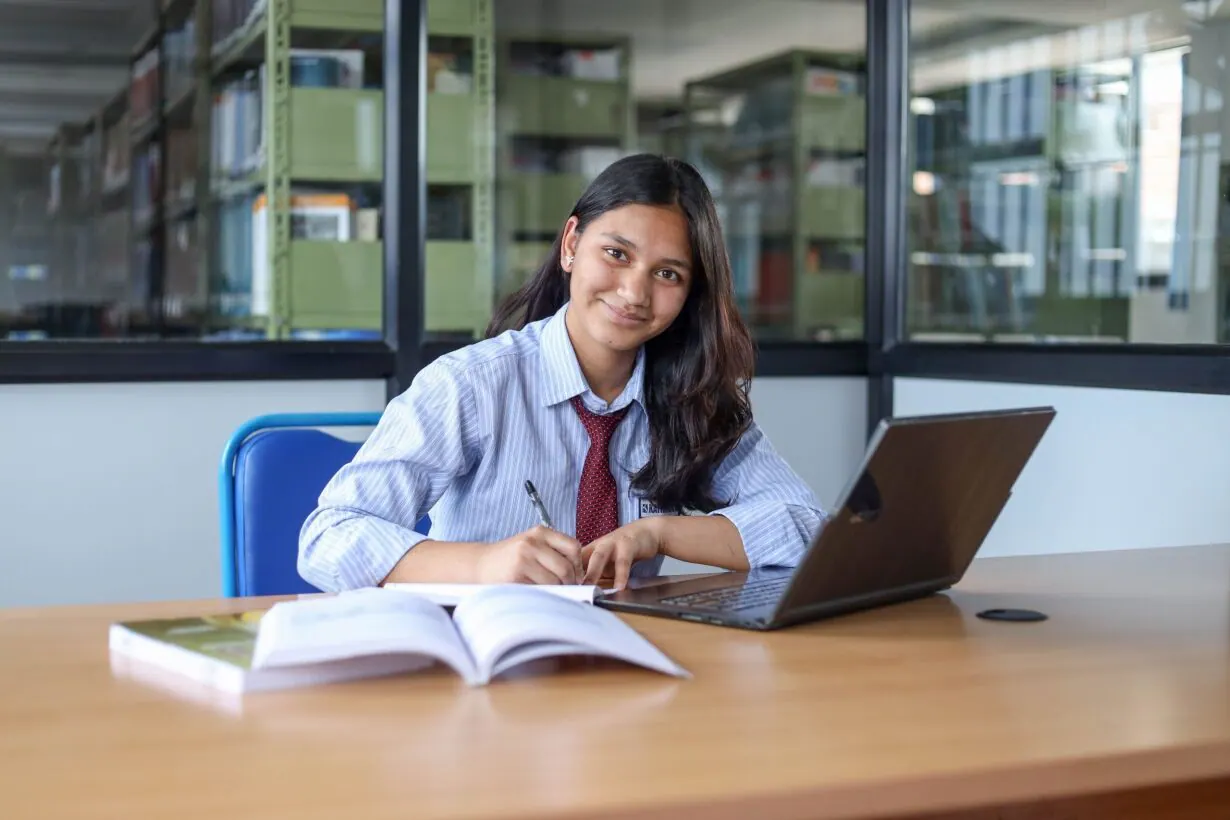 A student working at a laptop and an open book at a library table, stacks behind her