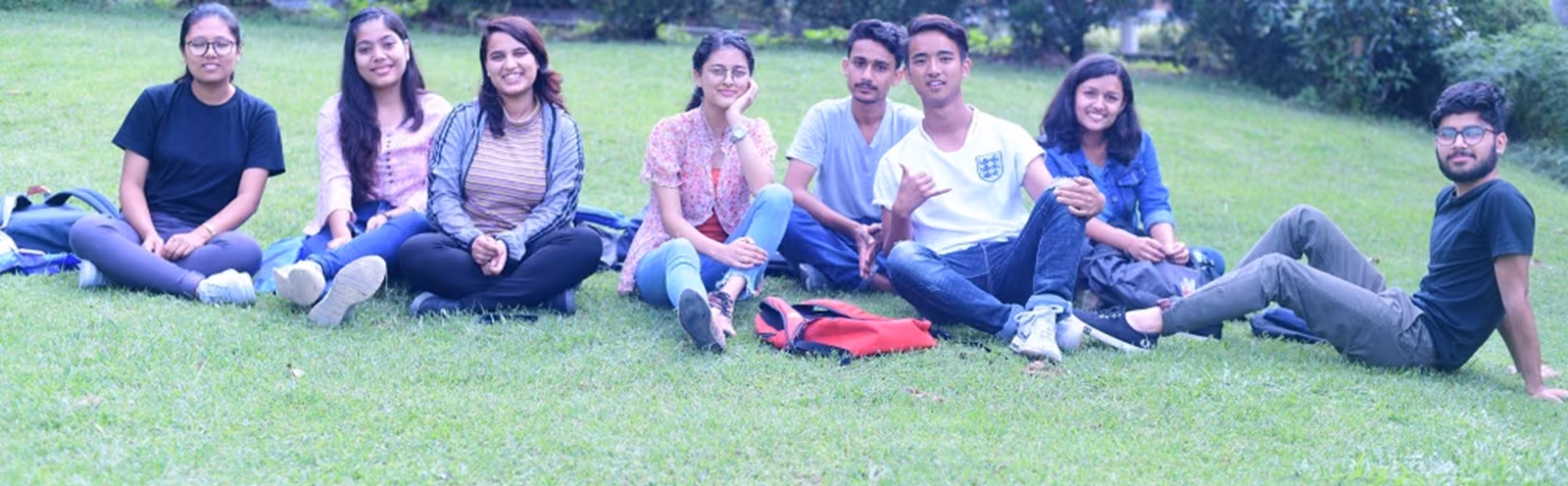 Eight students sitting together on the lawn, a hedge and trees behind them