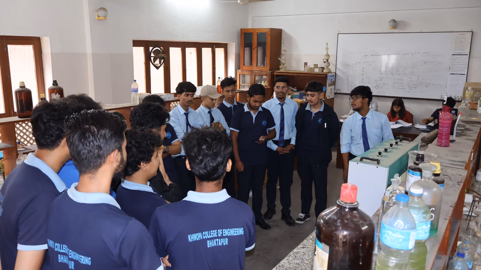 Students in college shirts round a laboratory bench of reagent bottles and burners
