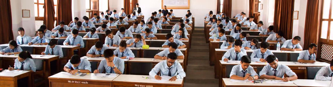Students at single desks through a long examination hall, curtains drawn along the windows
