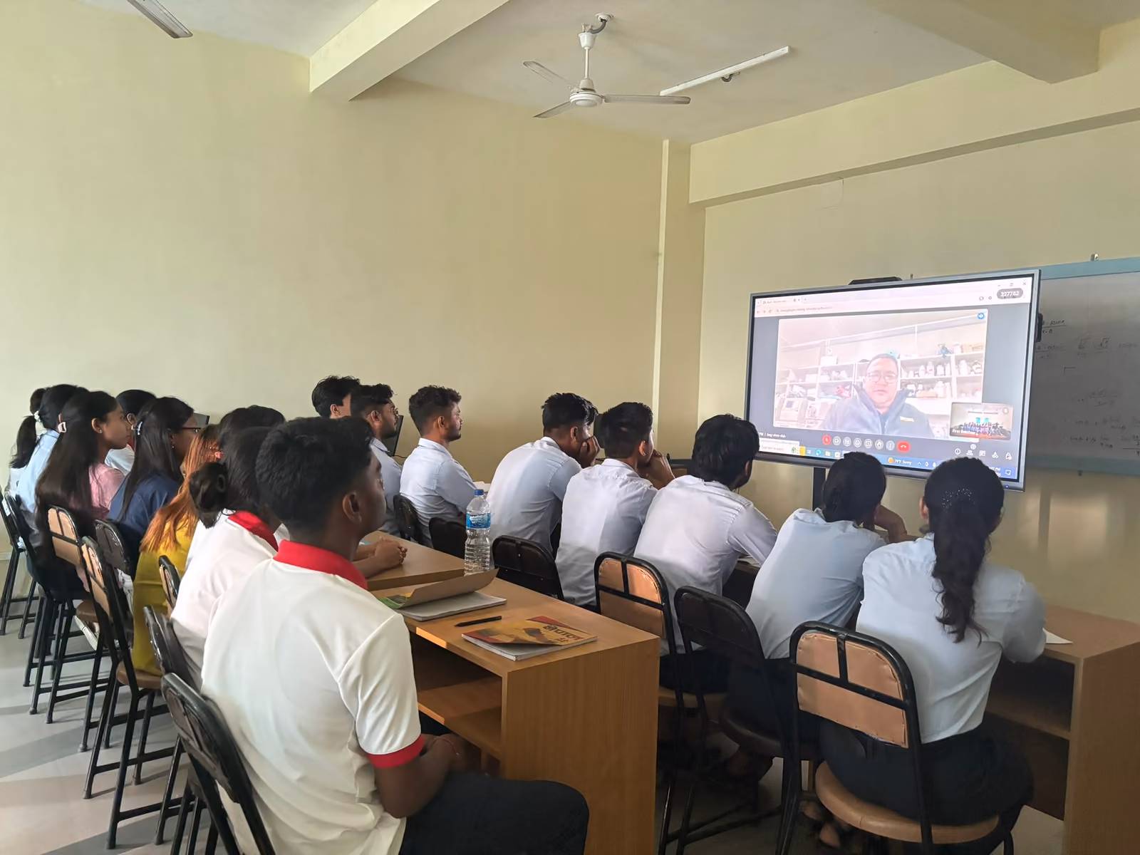 Students seated in a bright classroom facing a wall-mounted screen at the front of the room