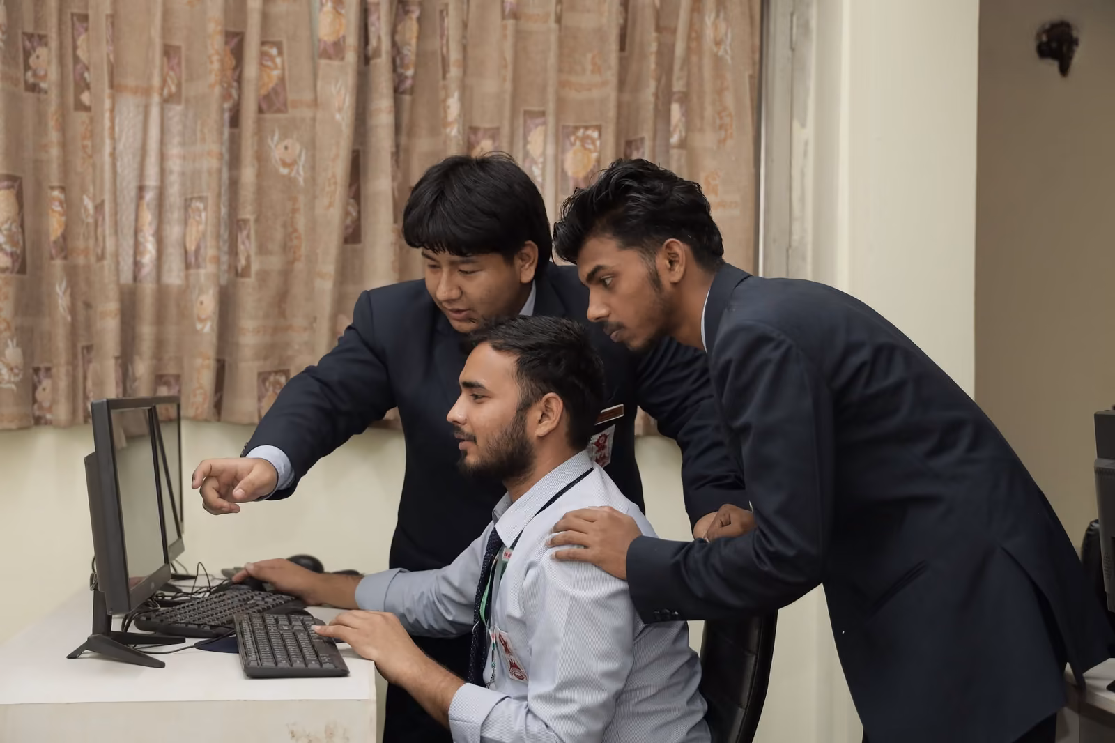 Two students leaning over a third at a desktop computer, pointing at the screen