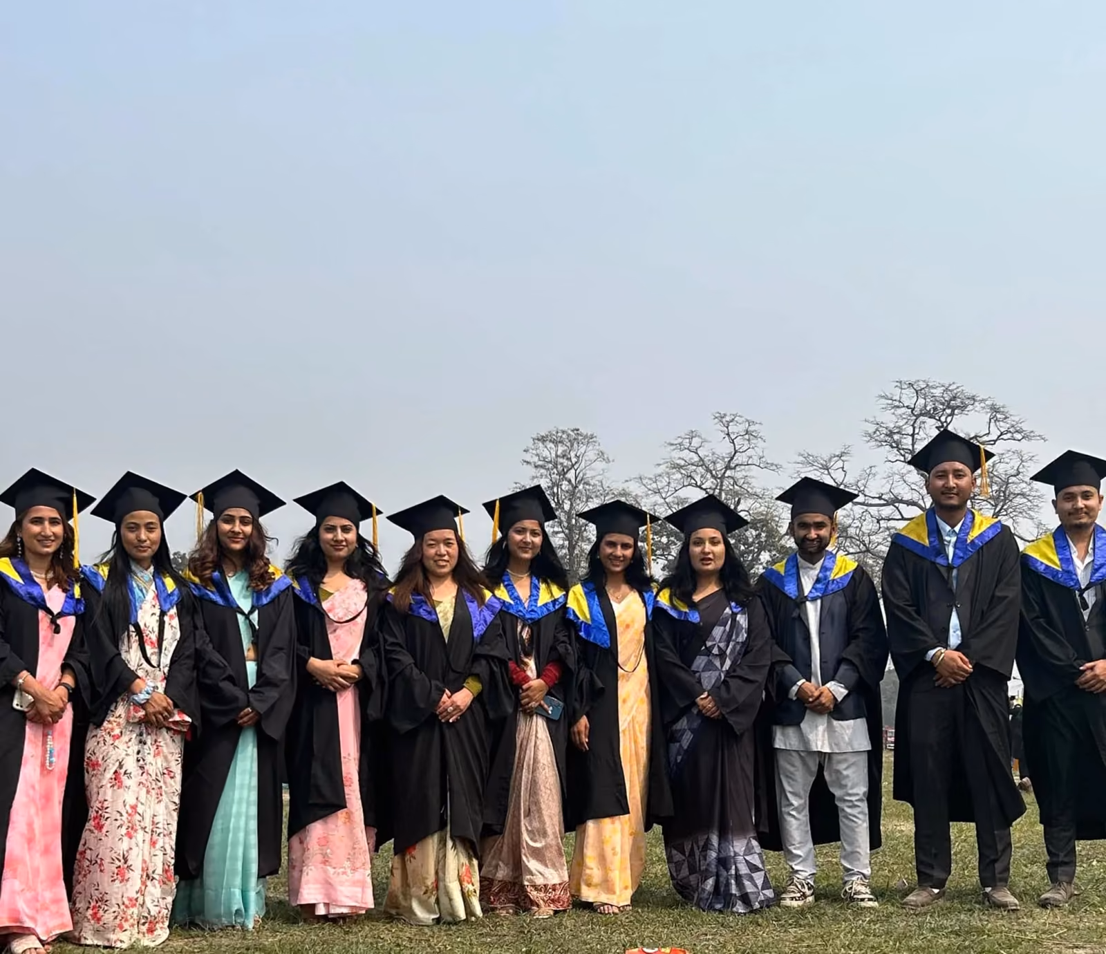 Graduates in caps and gowns lined up on the grass under a bare tree