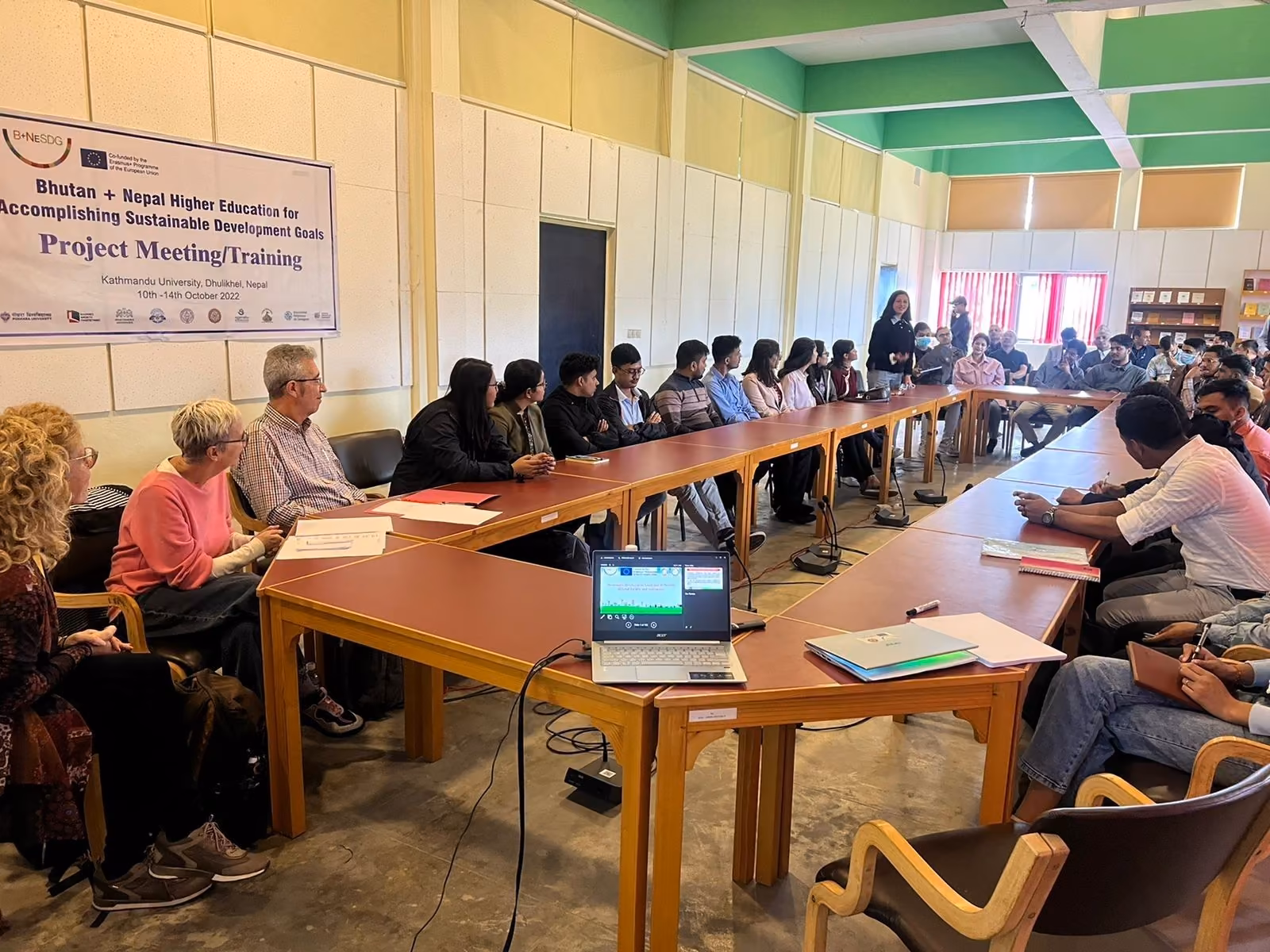 Participants round a long conference table under a project meeting banner, laptops open