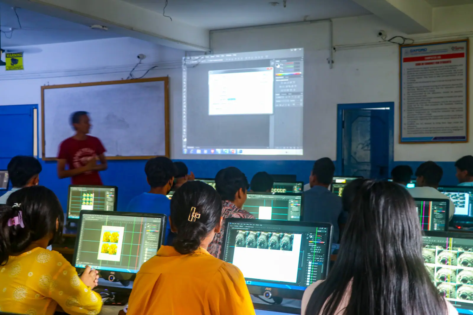 Students at desktop computers in a darkened laboratory, a presenter beside the projected screen