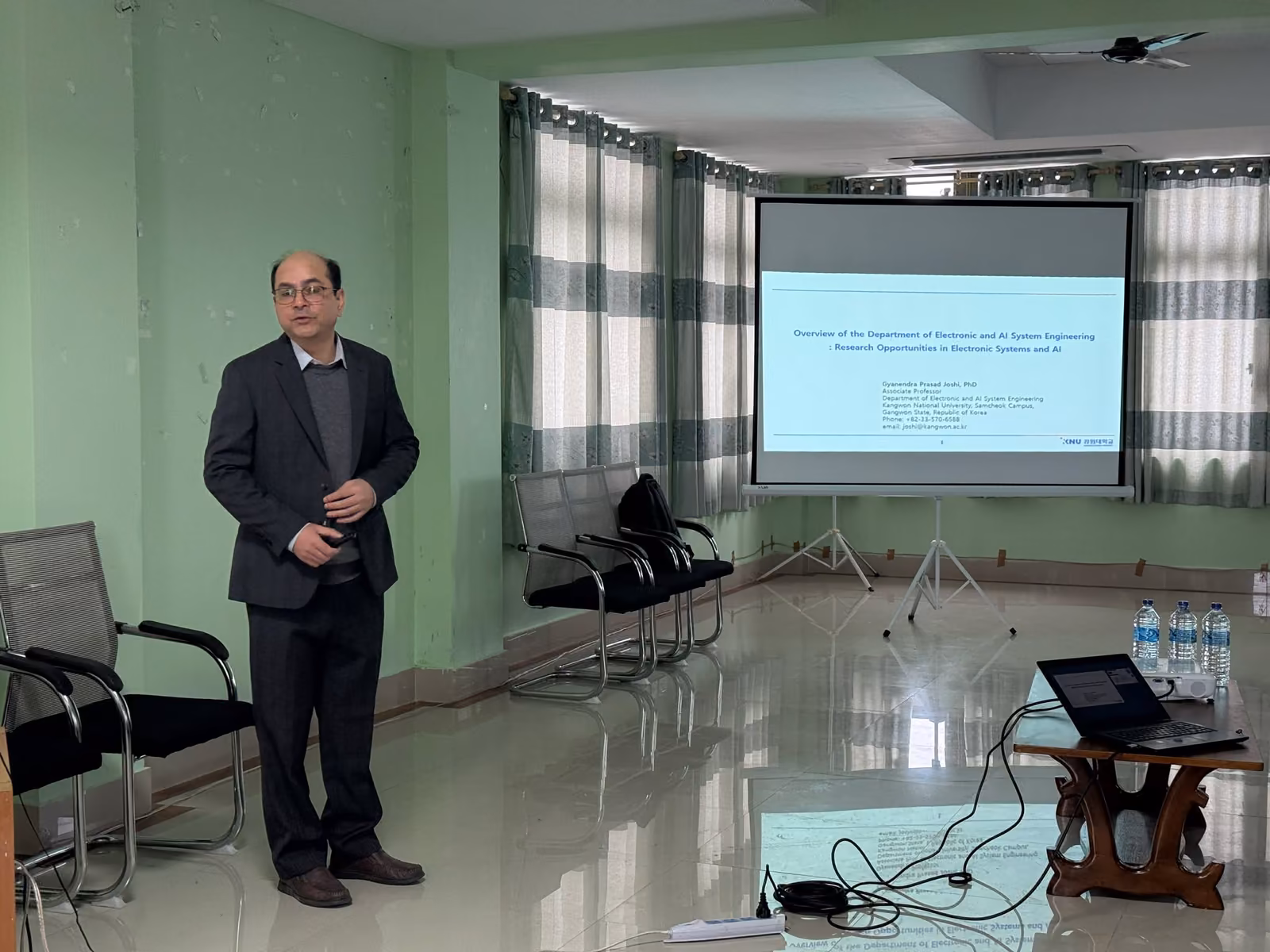A speaker beside a projected slide in the seminar hall, rows of chairs waiting to be filled