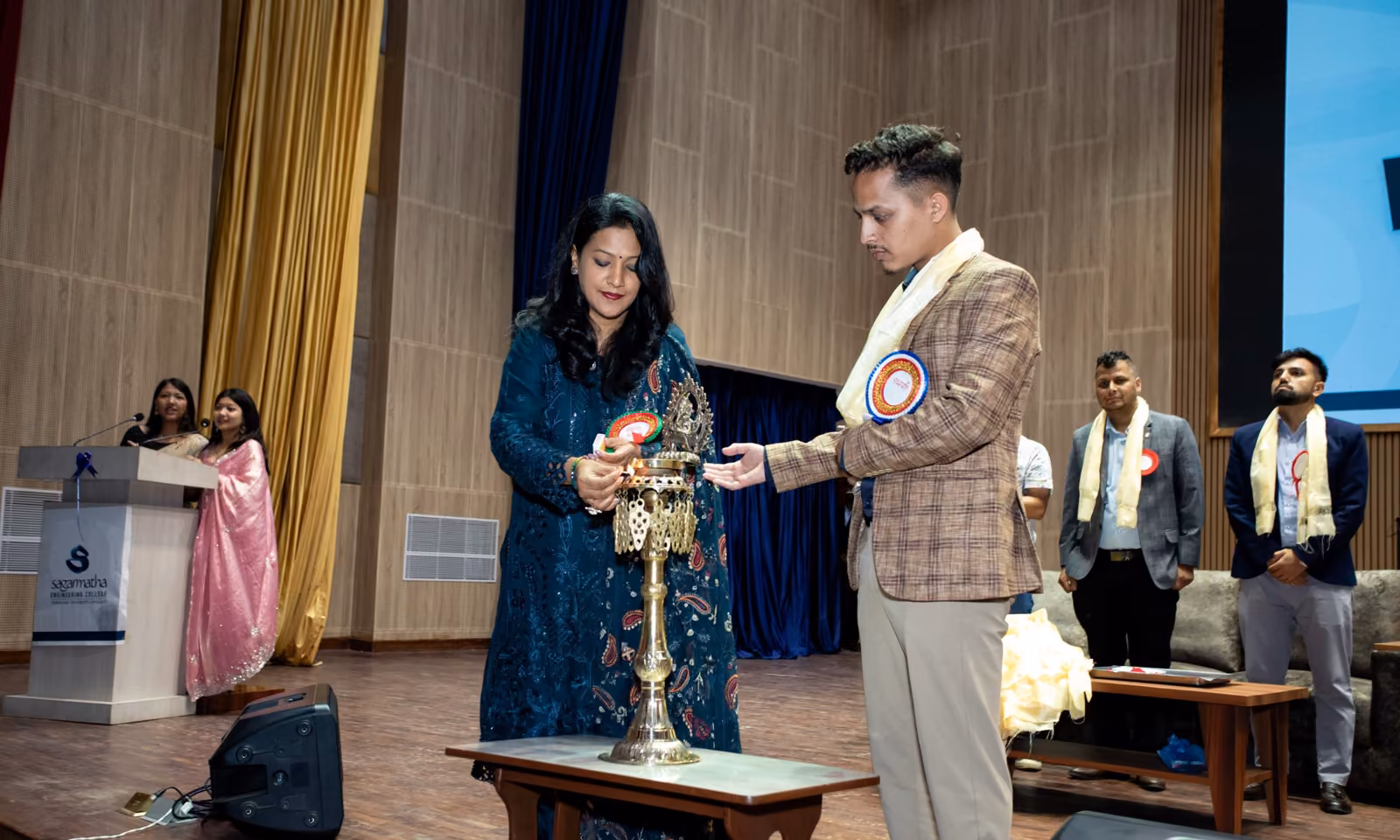A lamp being lit on stage at a college programme, guests waiting beside it
